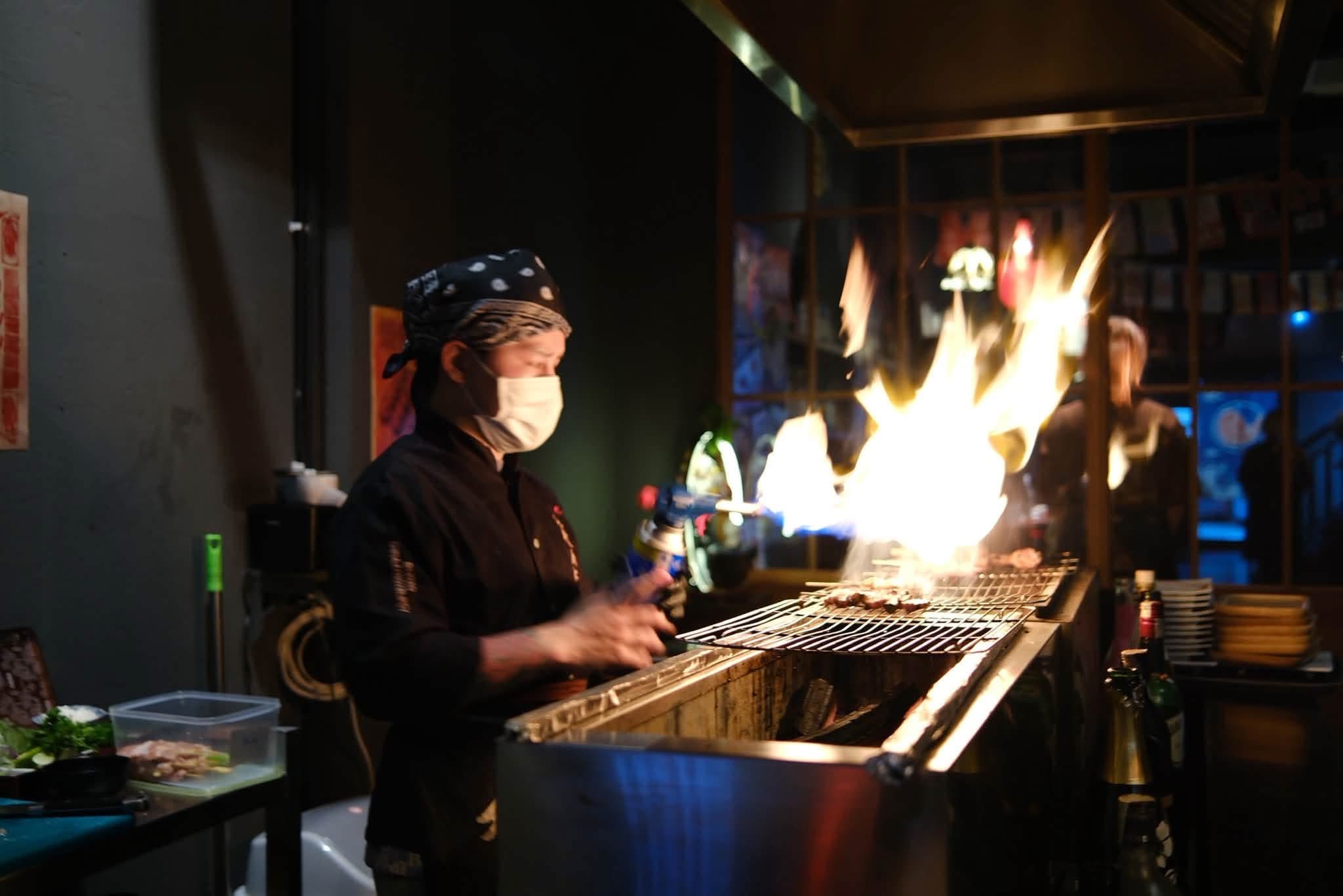 Đầu bếp EN Izakaya nướng xiên trên bếp than, lửa bùng lên trong quán tối